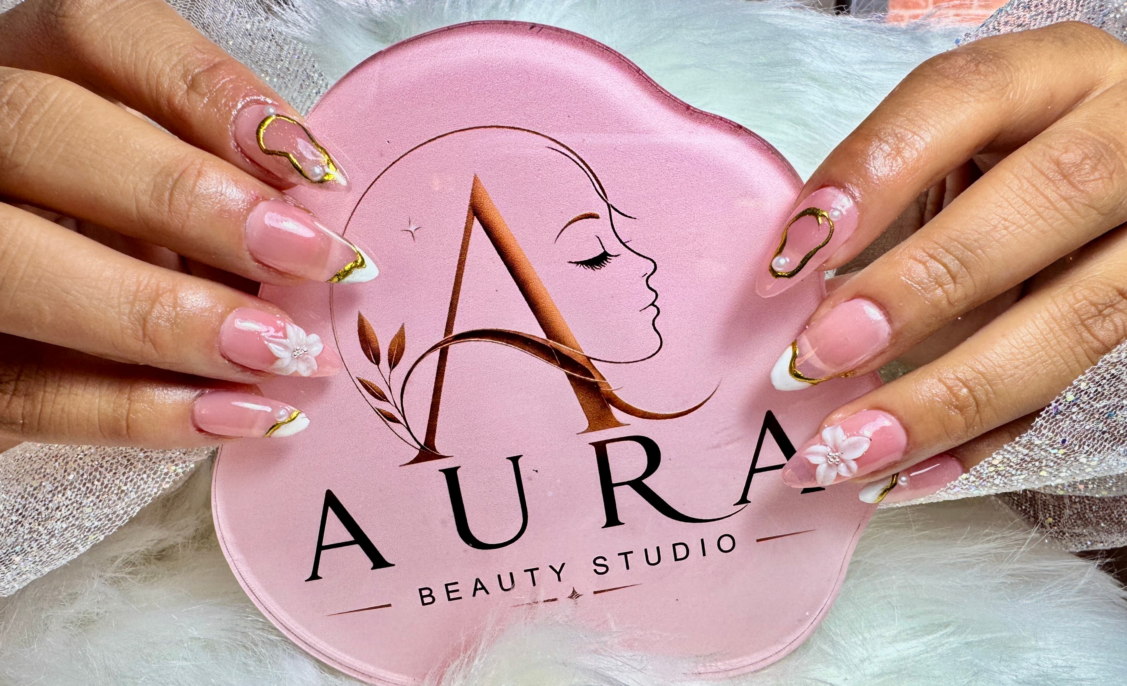 Manos con uñas almendradas nude, french, flores 3D y acentos dorados, sosteniendo la paleta con el logo de Aura Beauty Studio