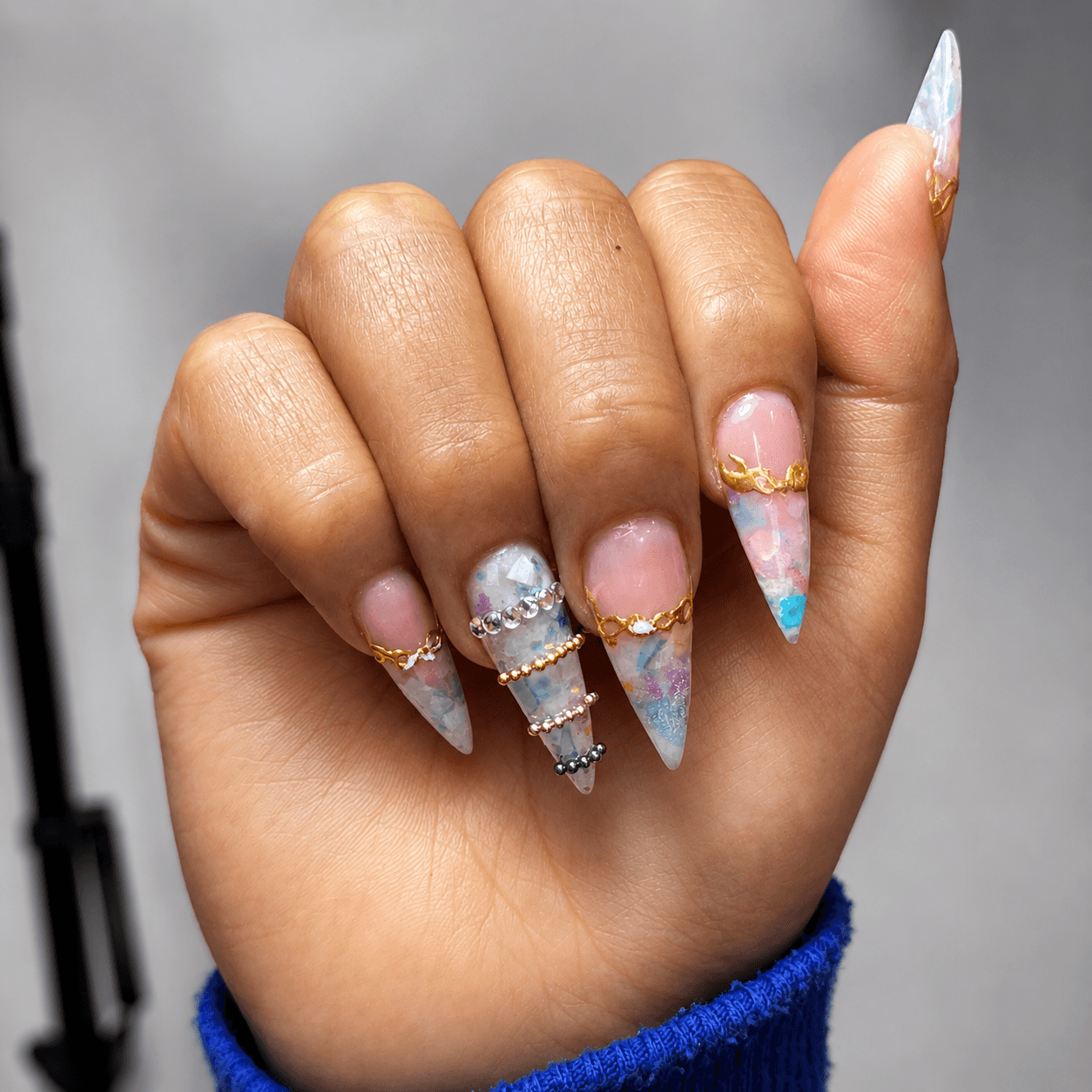 Mano con uñas stiletto encapsuladas en tonos pastel, cristales y detalles dorados