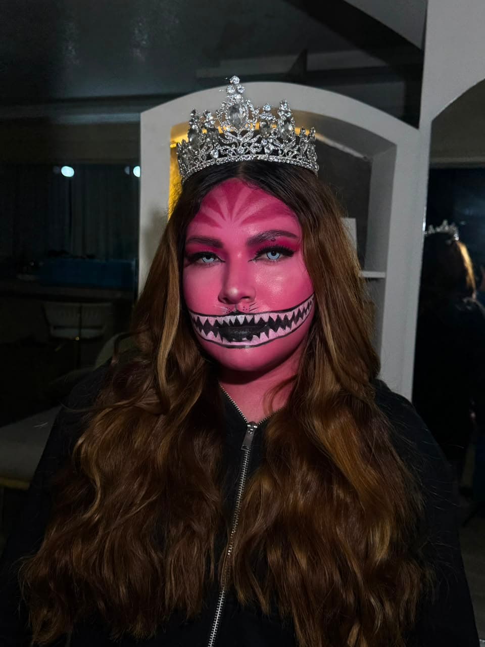 Maquillaje artístico de fantasía inspirado en el Gato de Cheshire, con rostro en rosa, sonrisa dentada pintada y corona de cristales