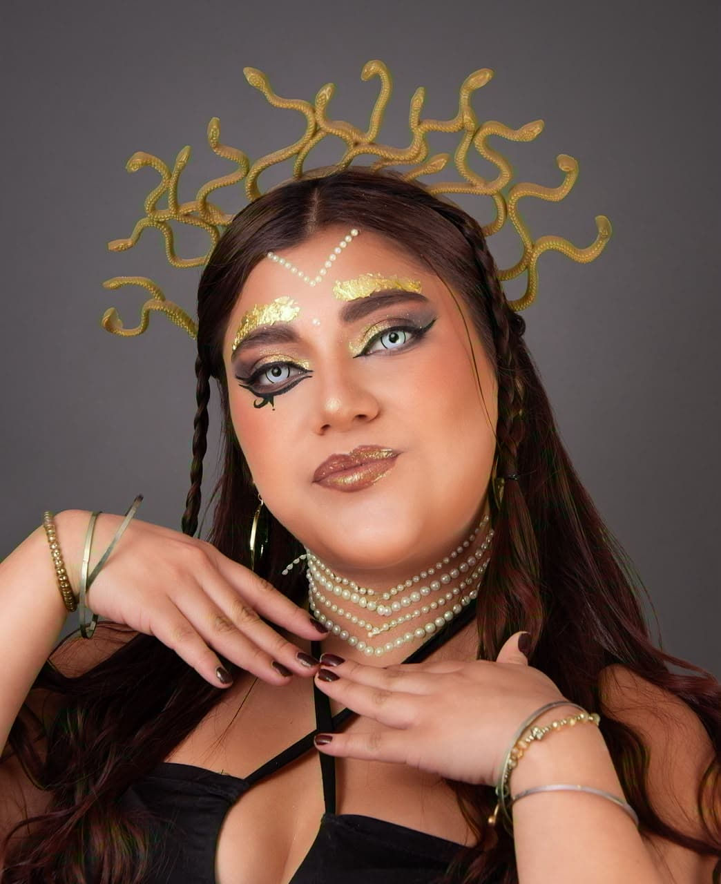 Maquillaje artístico de Medusa con delineado gráfico, pan de oro en cejas y labios, tocado de serpientes doradas y accesorios de perlas
