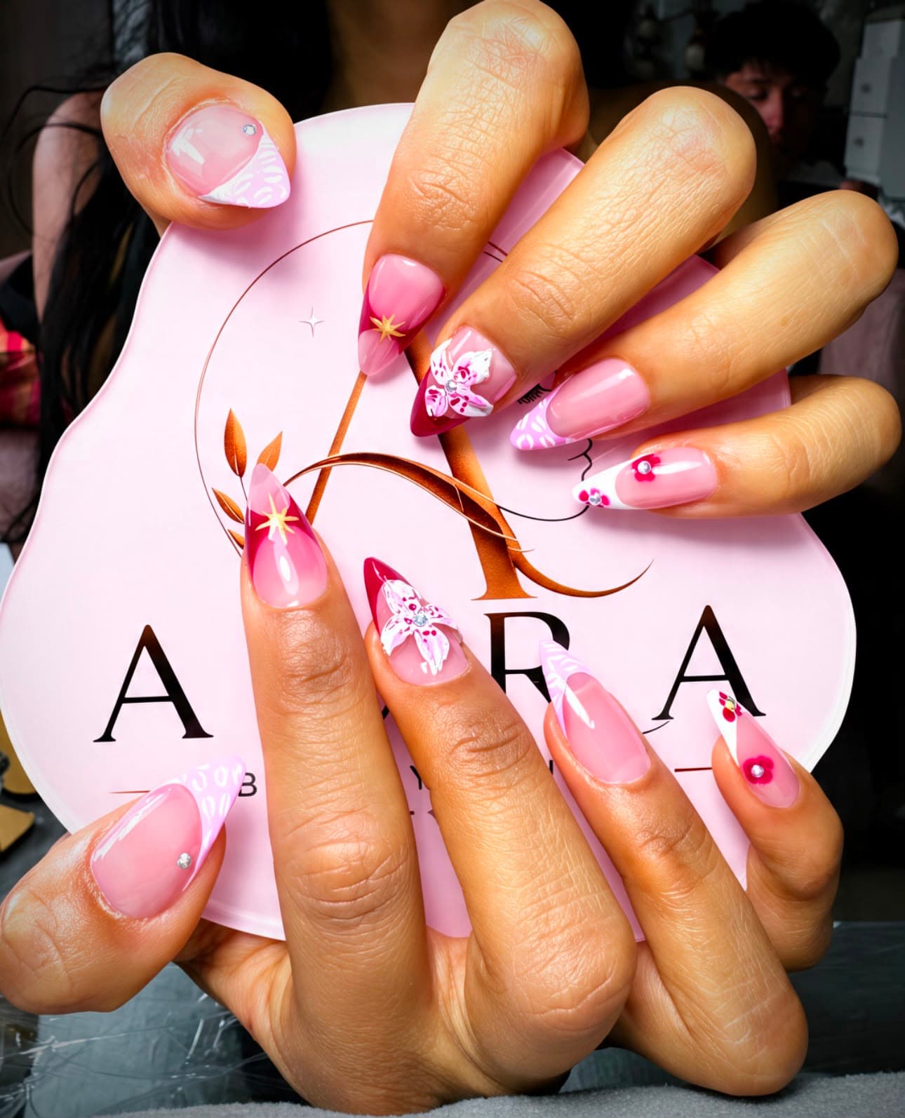 Manos con uñas almendradas en baby boomer rosa, decoradas con mariposas 3D, flores rojas y cristales, sosteniendo la paleta del logo de Aura Beauty Studio