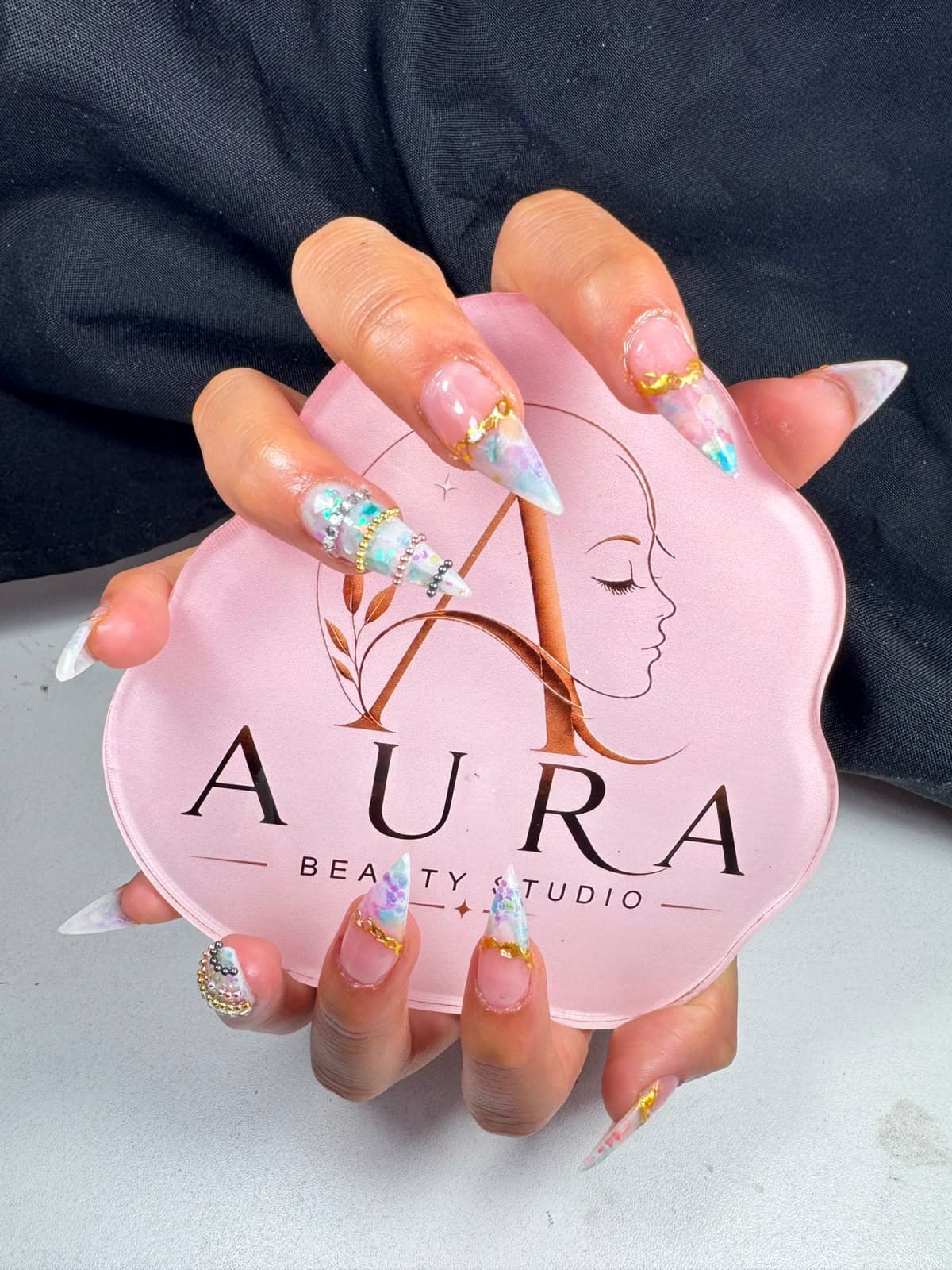 Uñas acrílicas largas en punta stiletto con efecto encapsulado nacarado, detalles en foil dorado y perlas, sosteniendo la paleta rosa con el logo de Aura Beauty Studio